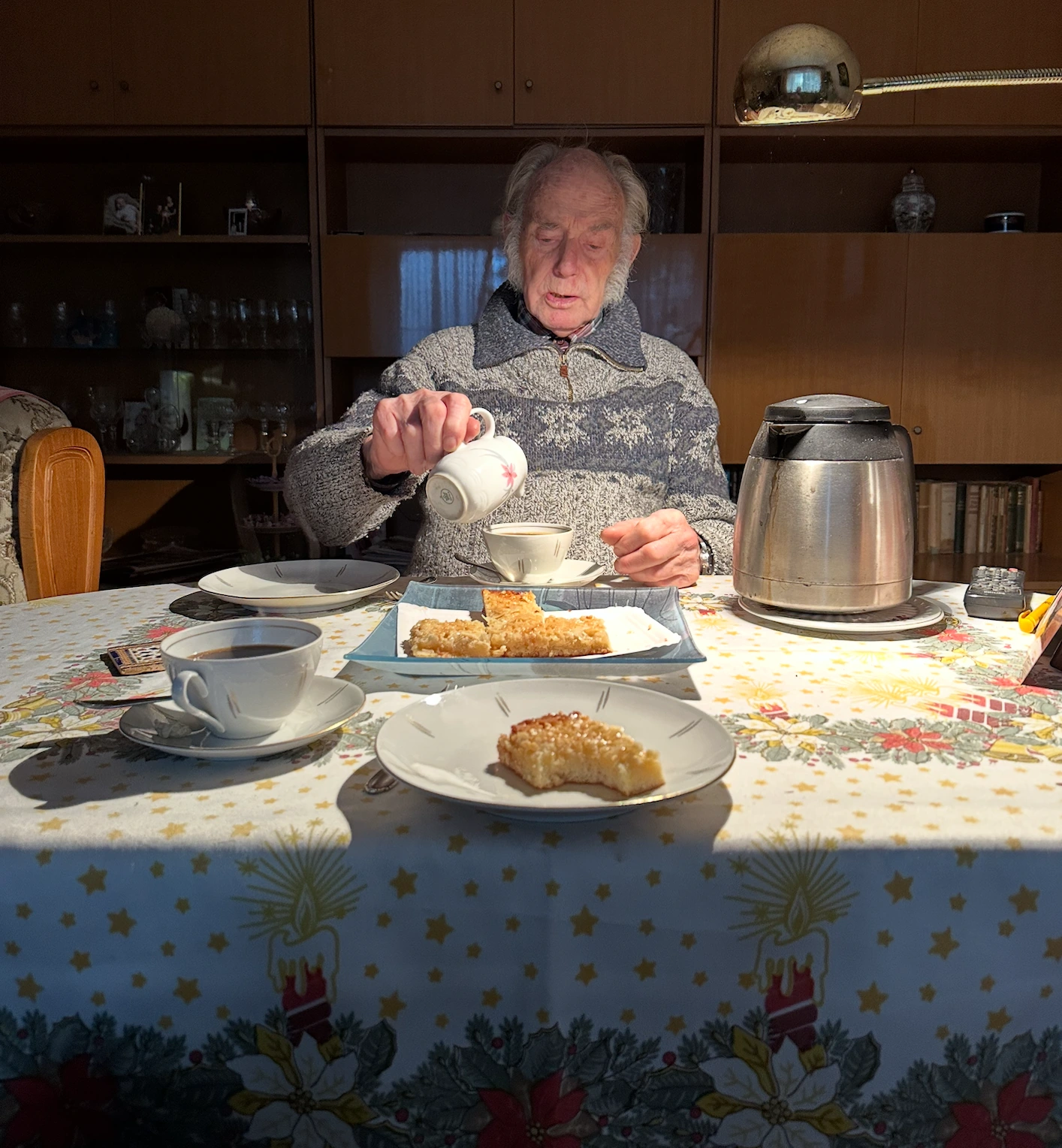 Michaels Opa am Tisch mit Kaffeetassen, Kuchen und einer kleinen Kanne in der Hand.