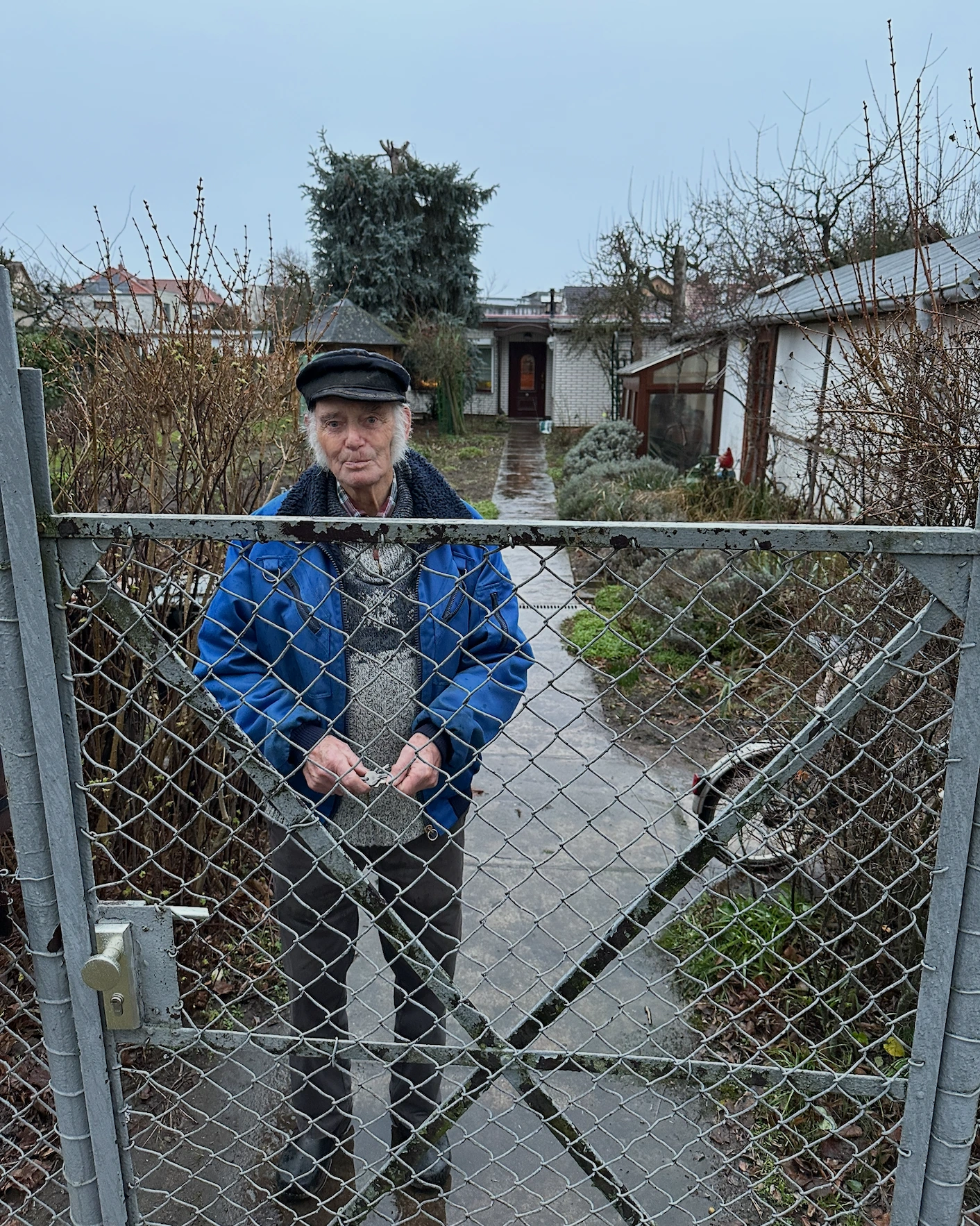 Michaels Opa steht mit blauer Jacke und dunkler Mütze am Gartentor.